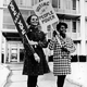Two smiling women stand outdoors and hold signs reading ‘Vote Baby Vote’ and ‘Voting is People Power,’ c. 1970.